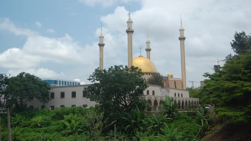 The Abuja National Mosque with its golden dome and four tall minarets, seen above trees and green vegetation under a cloudy sky