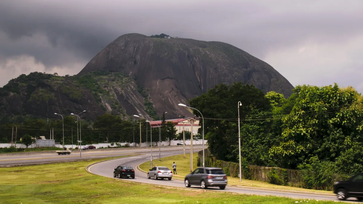 A large rounded rock face above trees and buildings, with cars on a curving road beneath a cloudy sky.
