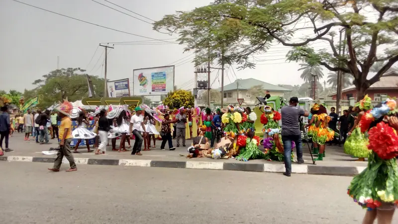 Carnival performers in colourful flower and leaf costumes dancing on a street in Calabar, watched by onlookers and a camera operator