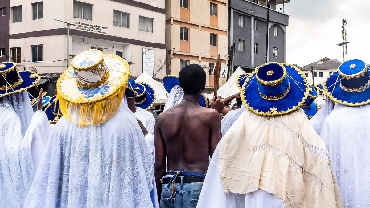 Eyo masquerade dancers seen from behind in flowing white robes and wide blue and gold hats, gathered on a Lagos street lined with buildings
