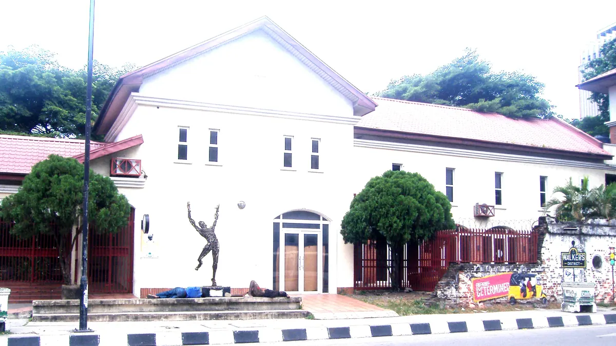 White two-storey building with a red tiled roof at Freedom Park, Lagos Island, with a bronze dancing figure statue in front and an old brick wall to the right