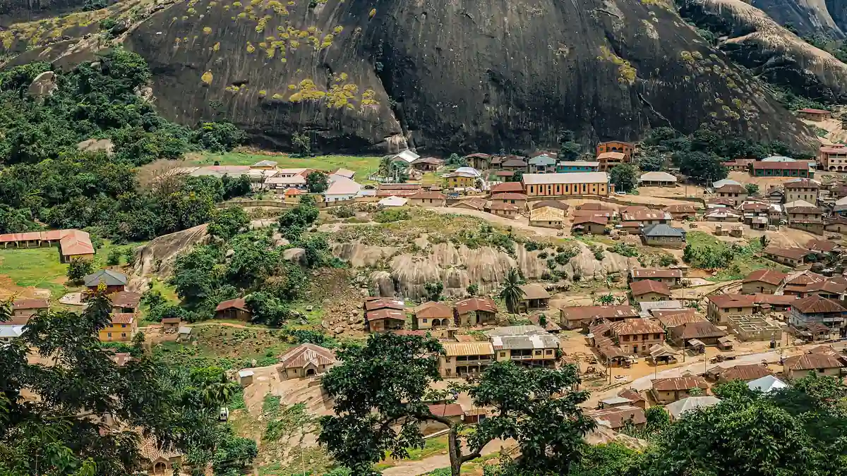 Large rounded rock faces of Idanre Hills above houses with metal roofs, trees and exposed rock in Ondo State.