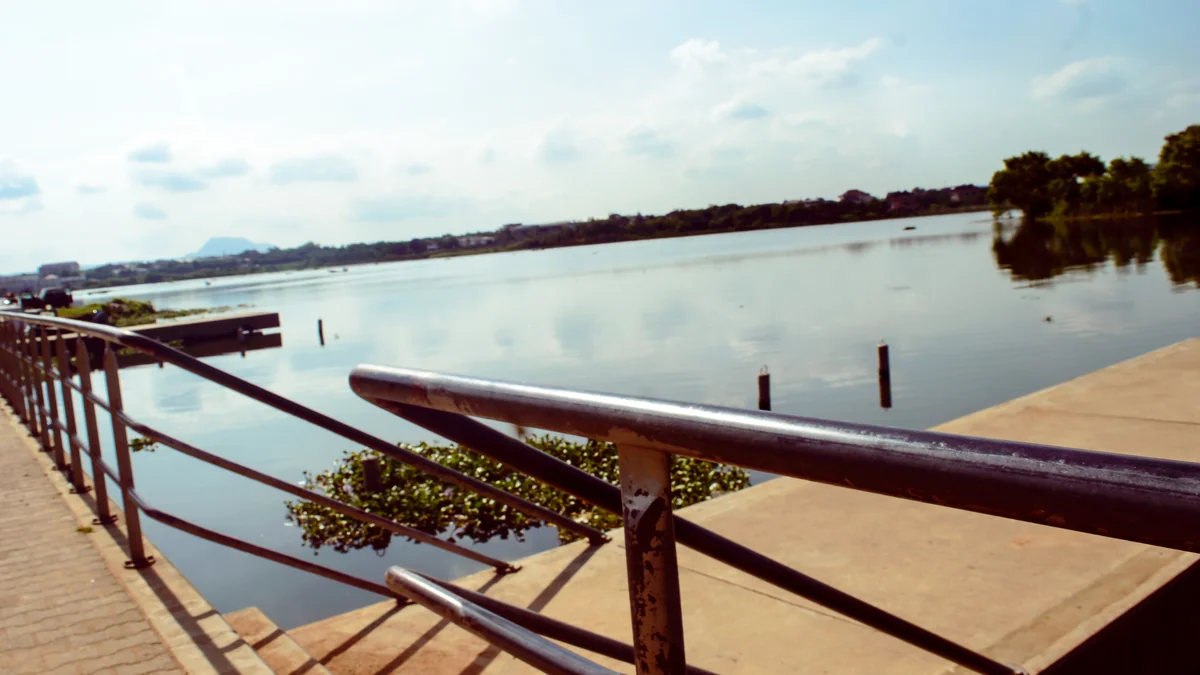 Calm water of Jabi Lake in Abuja seen from a paved lakeside walkway with metal railings, with trees and low hills on the far shore