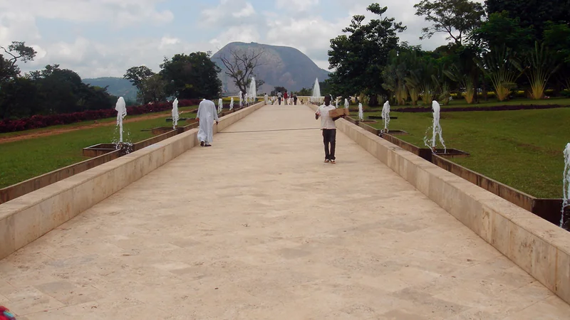A wide pale stone path lined with fountains and lawns at Millennium Park, Abuja, with walkers in the distance and Aso Rock rising behind the trees