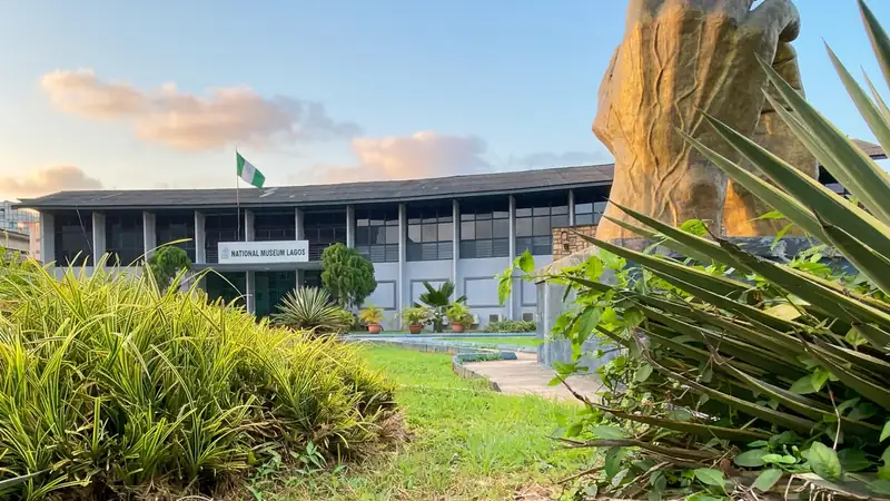 National Museum Lagos building behind a landscaped garden, with a large sculpted hand and spiky green plants in the foreground.