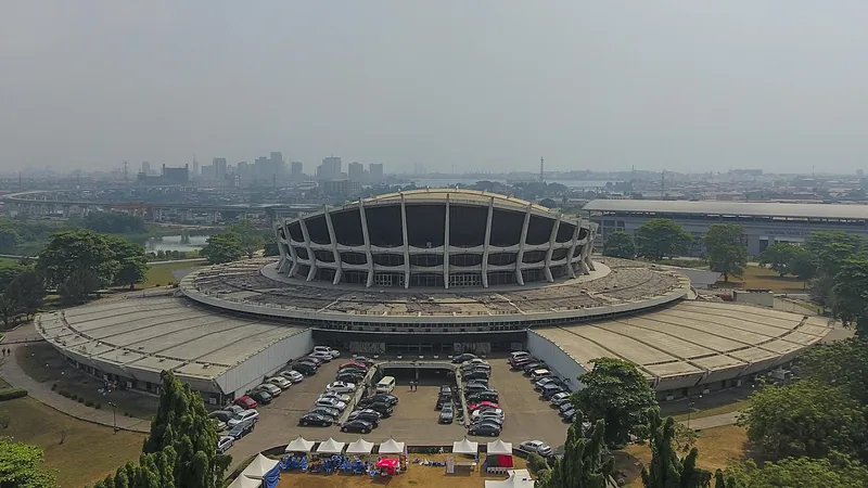Circular National Theatre building in Iganmu, Lagos, with sloping concrete supports, parked cars and the city skyline beyond.