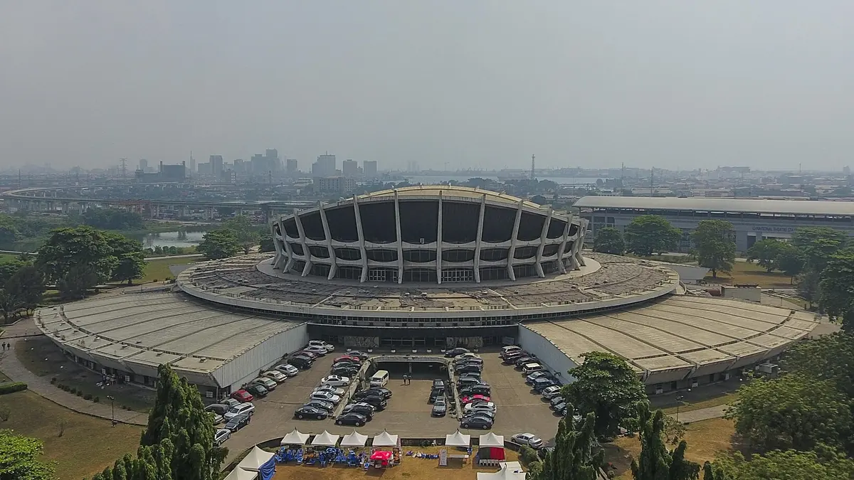 Circular National Theatre building in Iganmu, Lagos, with sloping concrete supports, parked cars and the city skyline beyond.