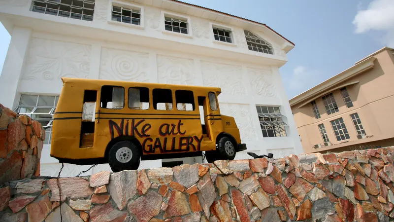 A yellow bus sculpture painted with the words Nike Art Gallery, mounted on a stone wall in front of the gallery's white building with carved wall panels
