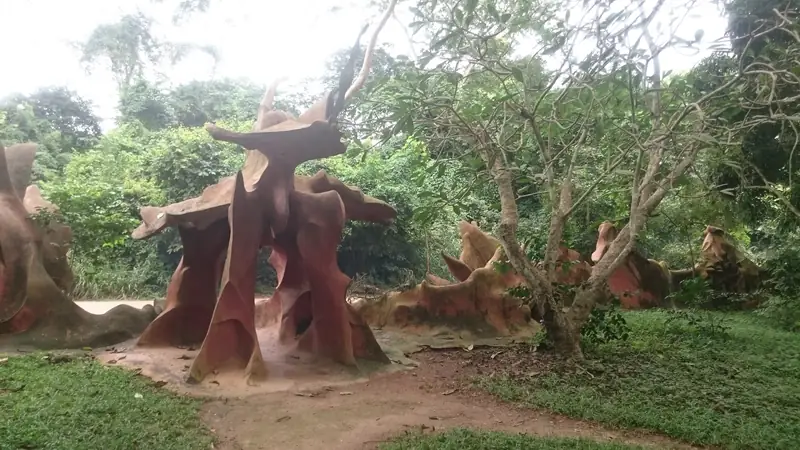 Reddish-brown sculptural forms beside a dirt path, surrounded by trees and green forest vegetation.