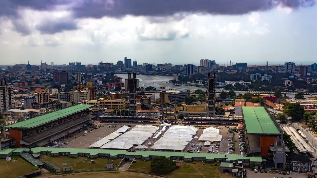 Aerial view of Tafawa Balewa Square on Lagos Island, with green-roofed stands around a large open ground covered in white tents, and the city skyline and lagoon beyond